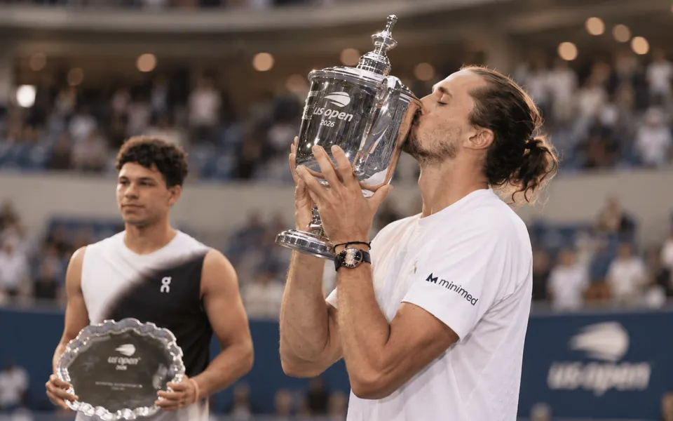 Alexander Zverev kisses the US Open trophy after winning the 2026 men’s final, with Ben Shelton holding the runner-up plate in the background.