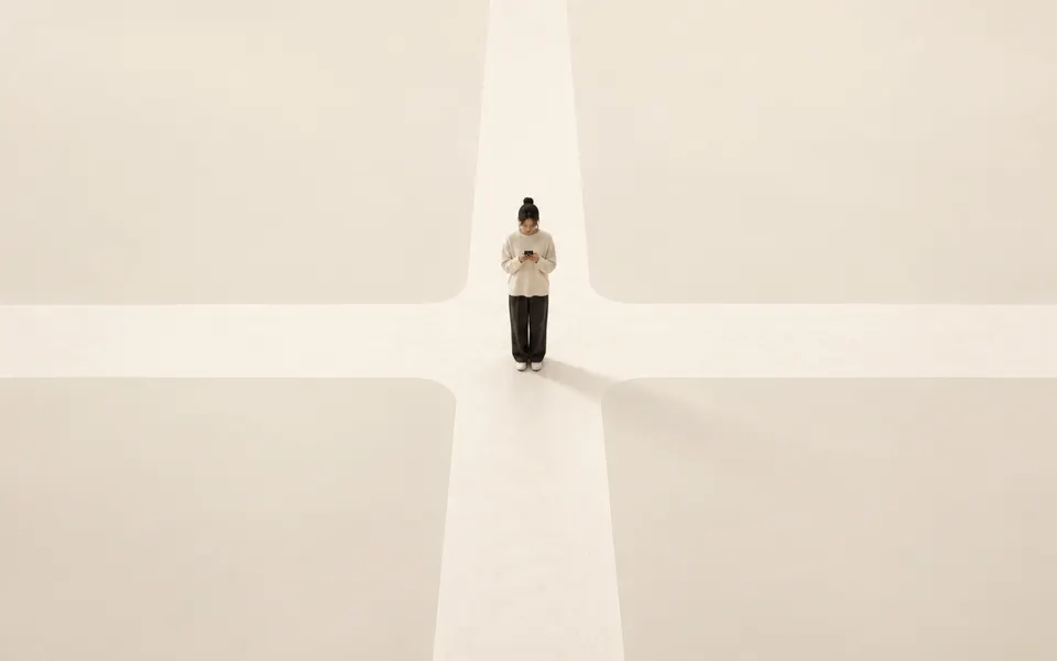 A person stands at the centre of an empty cream-coloured crossroads, looking down at a phone held in both hands.