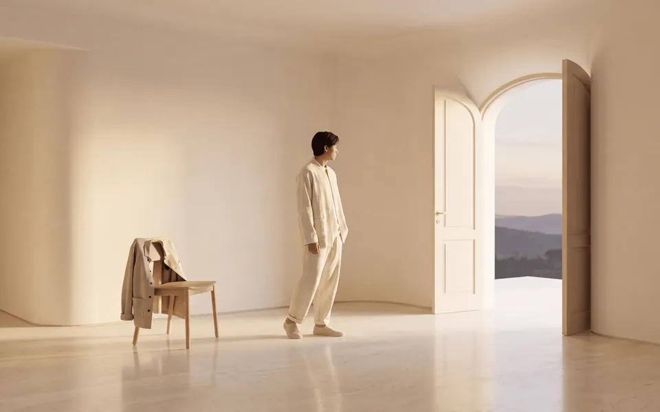 A person stands in a minimal cream room, looking toward an open doorway at dusk while a jacket rests on a chair behind them.