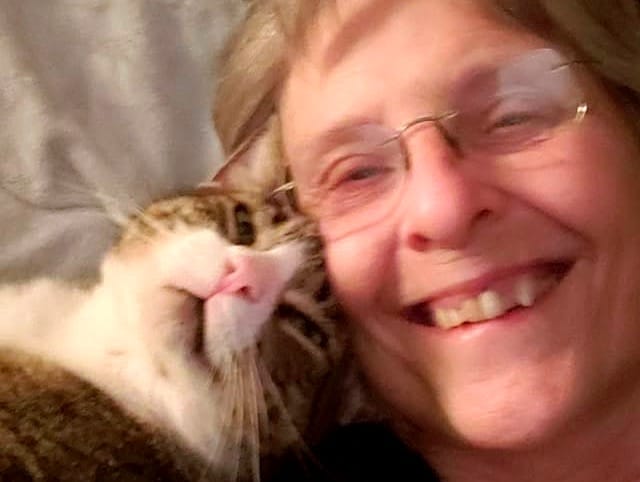 Closeup/selfie: Sweetie Boy’s handsome cat face looks into the camera, with the top of his head against Brooke’s right cheek while she also looks into the camera, smiling.