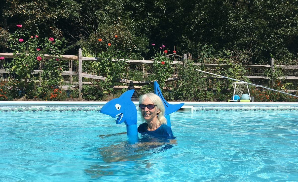 Janet is in a pool, wearing a blue t-shirt and floating with a foam shark. Her smile reveals her missing front tooth, and her bluejay earring matches the image's color palette 