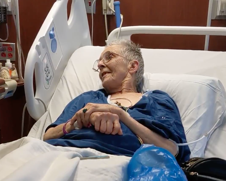 Brooke is sitting in a hospital bed during the second video described in this post. One hand is crossed over the other while she smiles speaking with a healthcare worker.