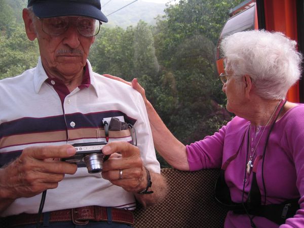Sitting in a gondola, Granddad reviews a photo on his digital camera while Grandmom rests a hand on his shoulder and looks out over the trees