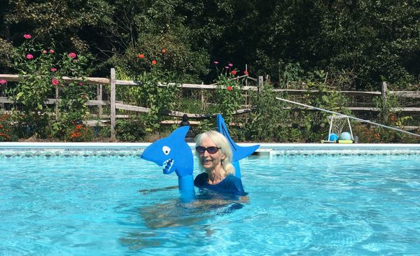 Janet is in a pool, wearing a blue t-shirt and floating with a foam shark. Her smile reveals her missing front tooth, and her bluejay earring matches the image's color palette 