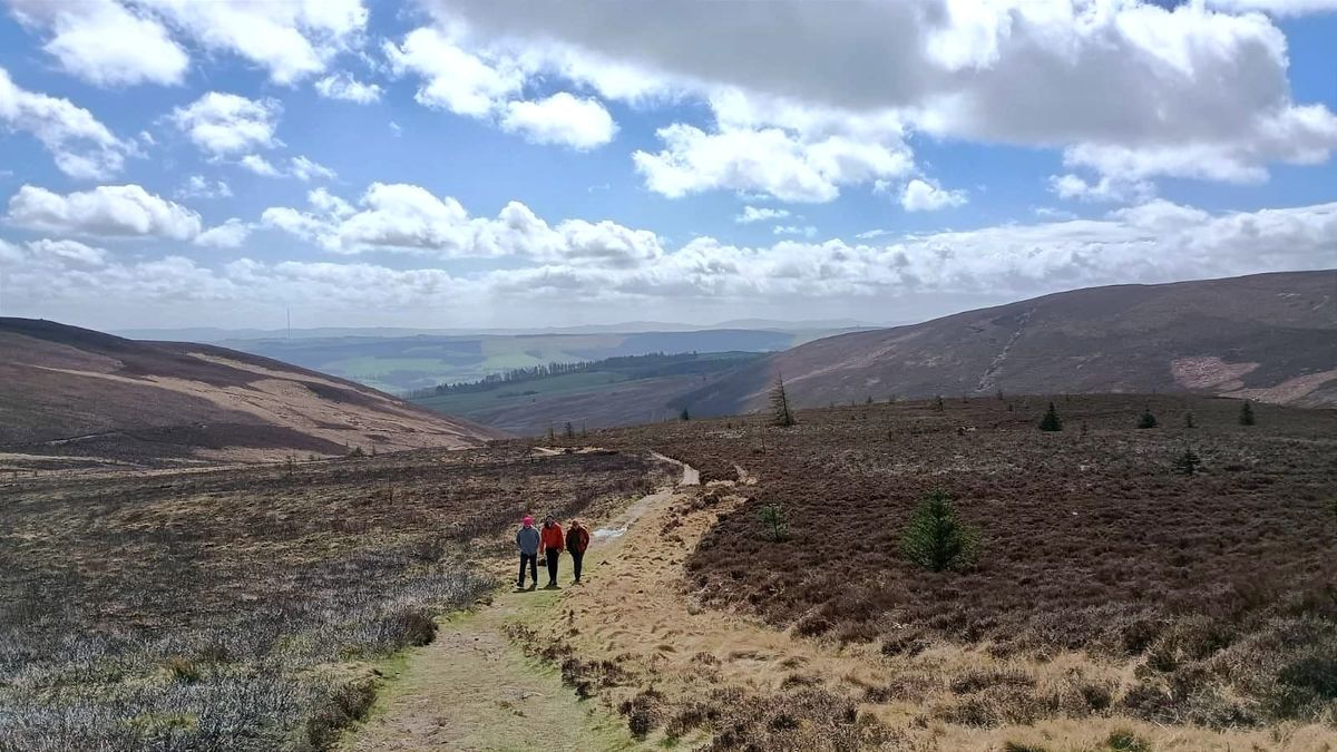 Three figures walking in the Scottish HIlls under a sky of sun and cloud