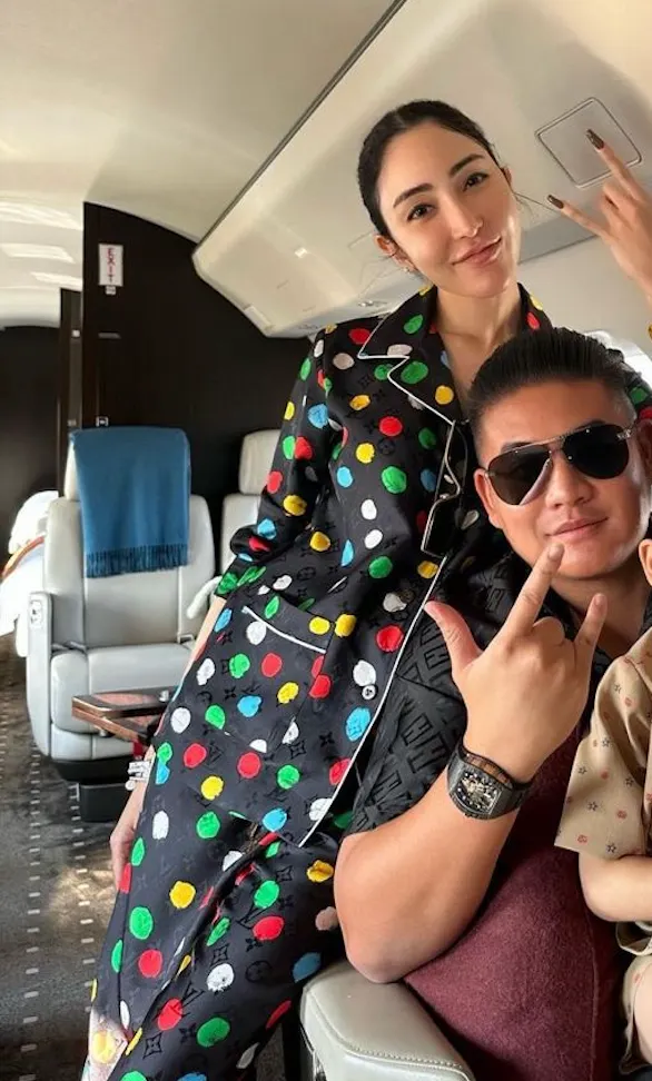Woman in colourful polka-dot pyjamas and man in sunglasses pull poses inside a private jet cabin