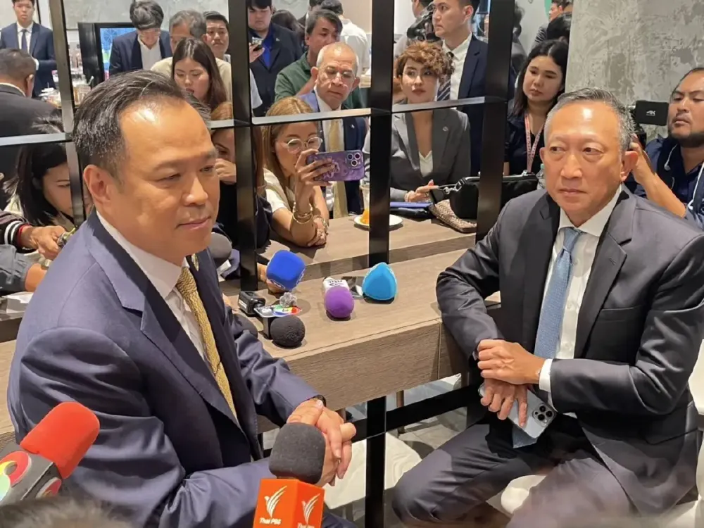 Two suited men sit at a table at a crowded press event, with microphones and reporters' phones raised around them