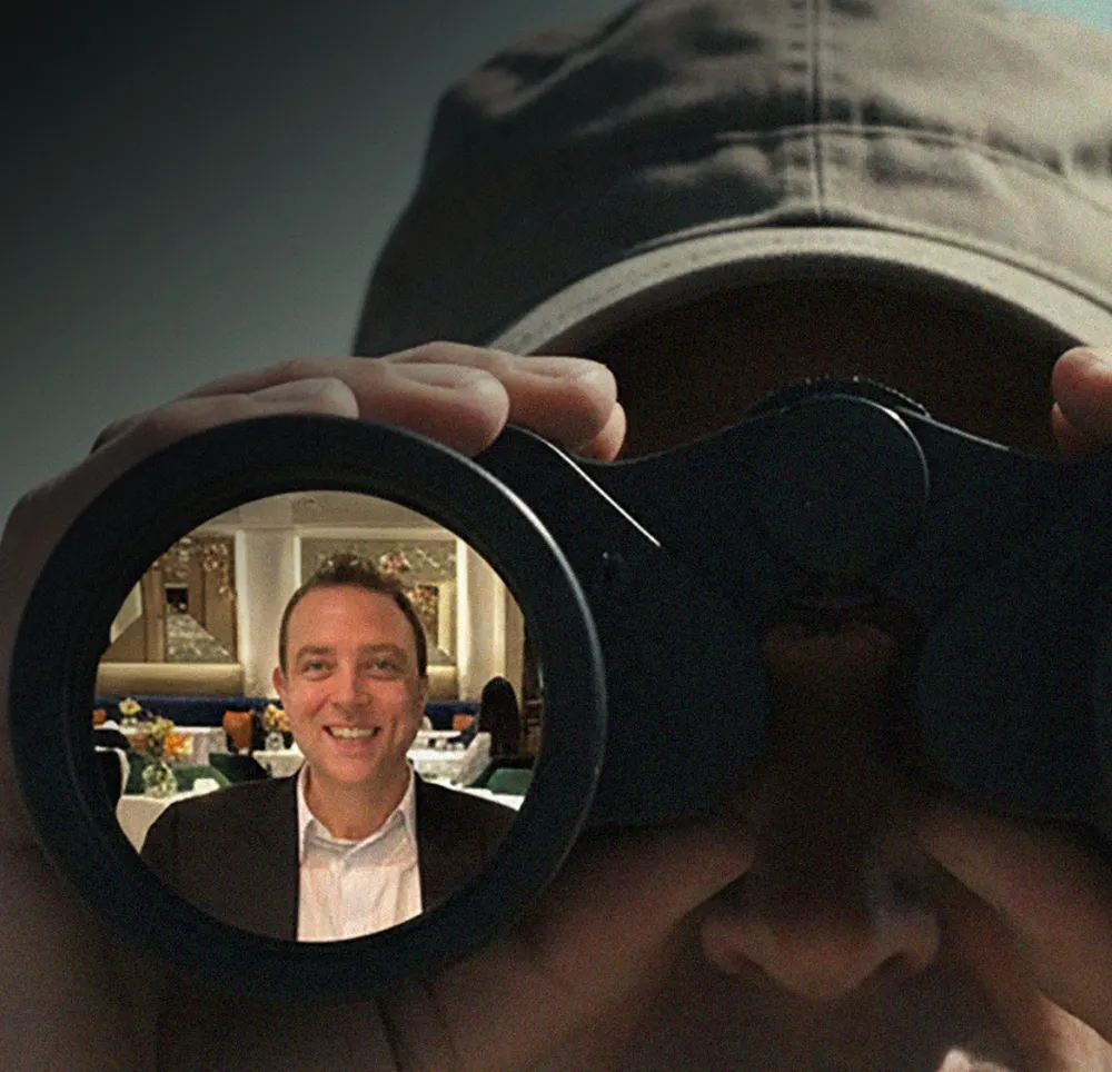 Man in a dark cap peers through binoculars; the lens shows a smiling man in a suit in a restaurant