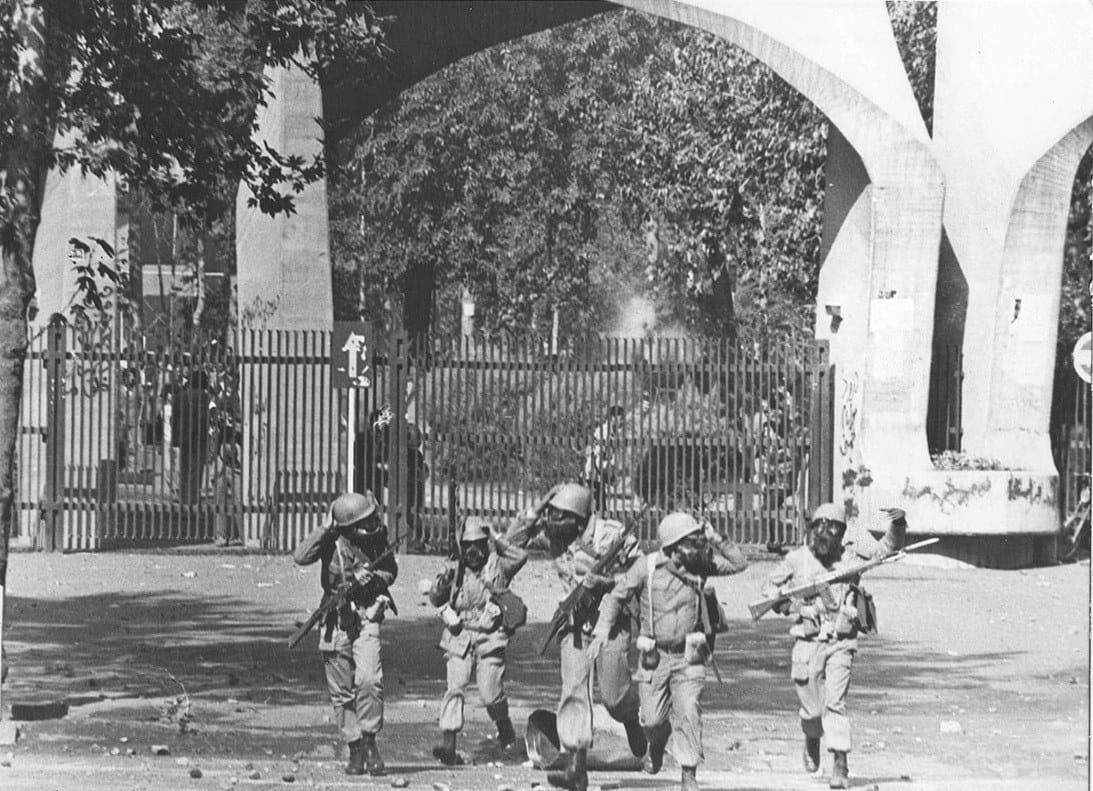 Five helmeted soldiers with rifles walk away from the arched main gate of Tehran University, stones scattered on the ground.