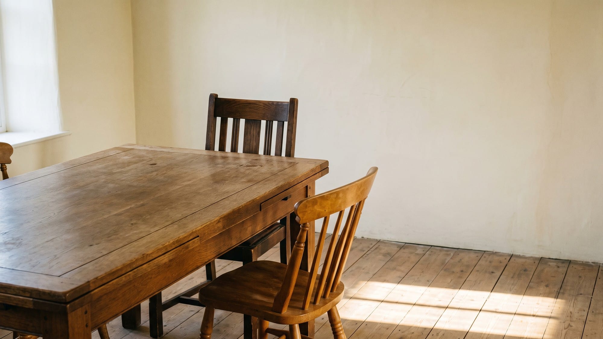 Two mismatched wooden chairs of different styles drawn up to the end of a long, empty oak table in a bare room, lit by low afternoon light across the floorboards.