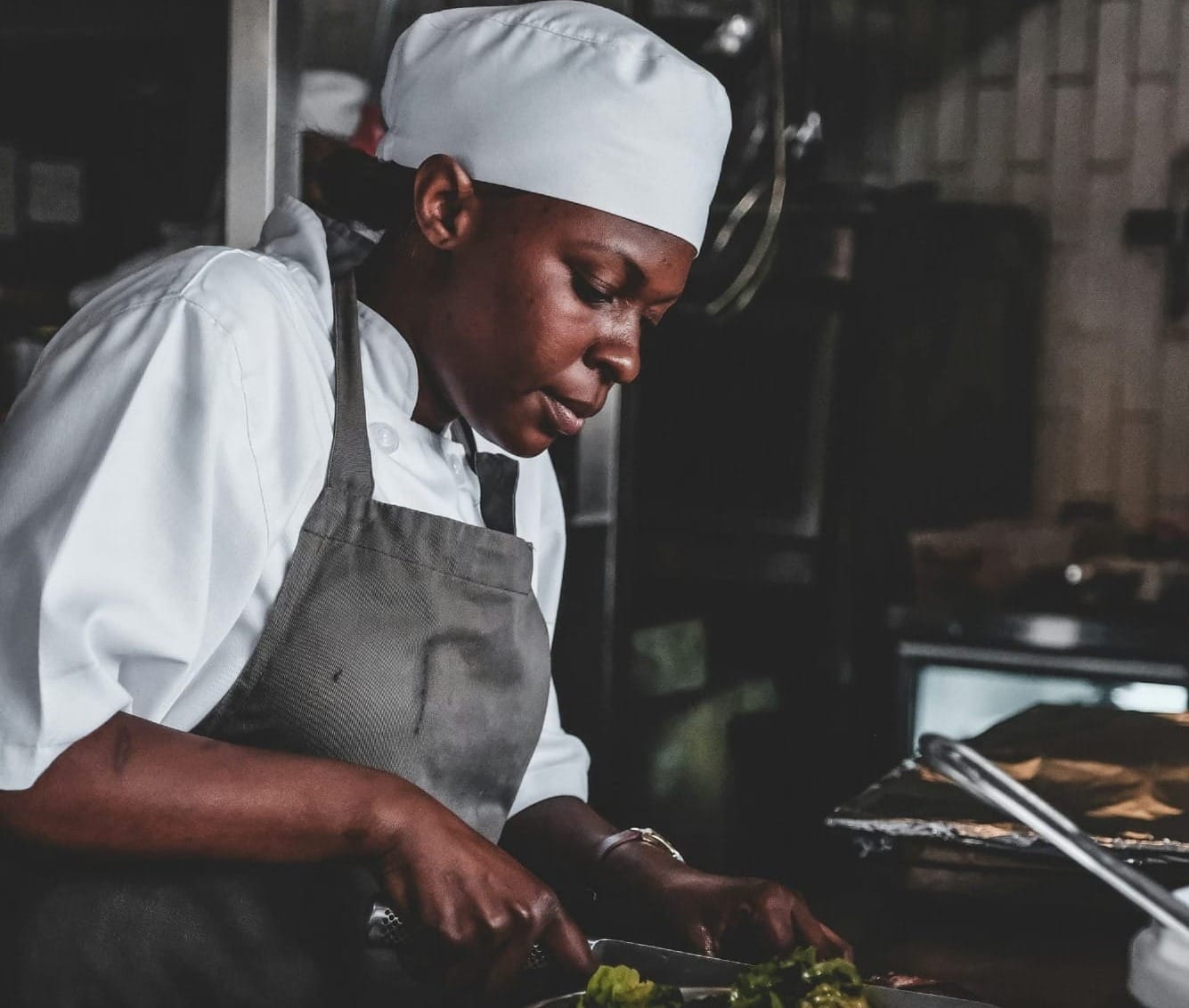 A chef in a white uniform and toque cuts ingredients over a bowl in a restaurant kitchen.