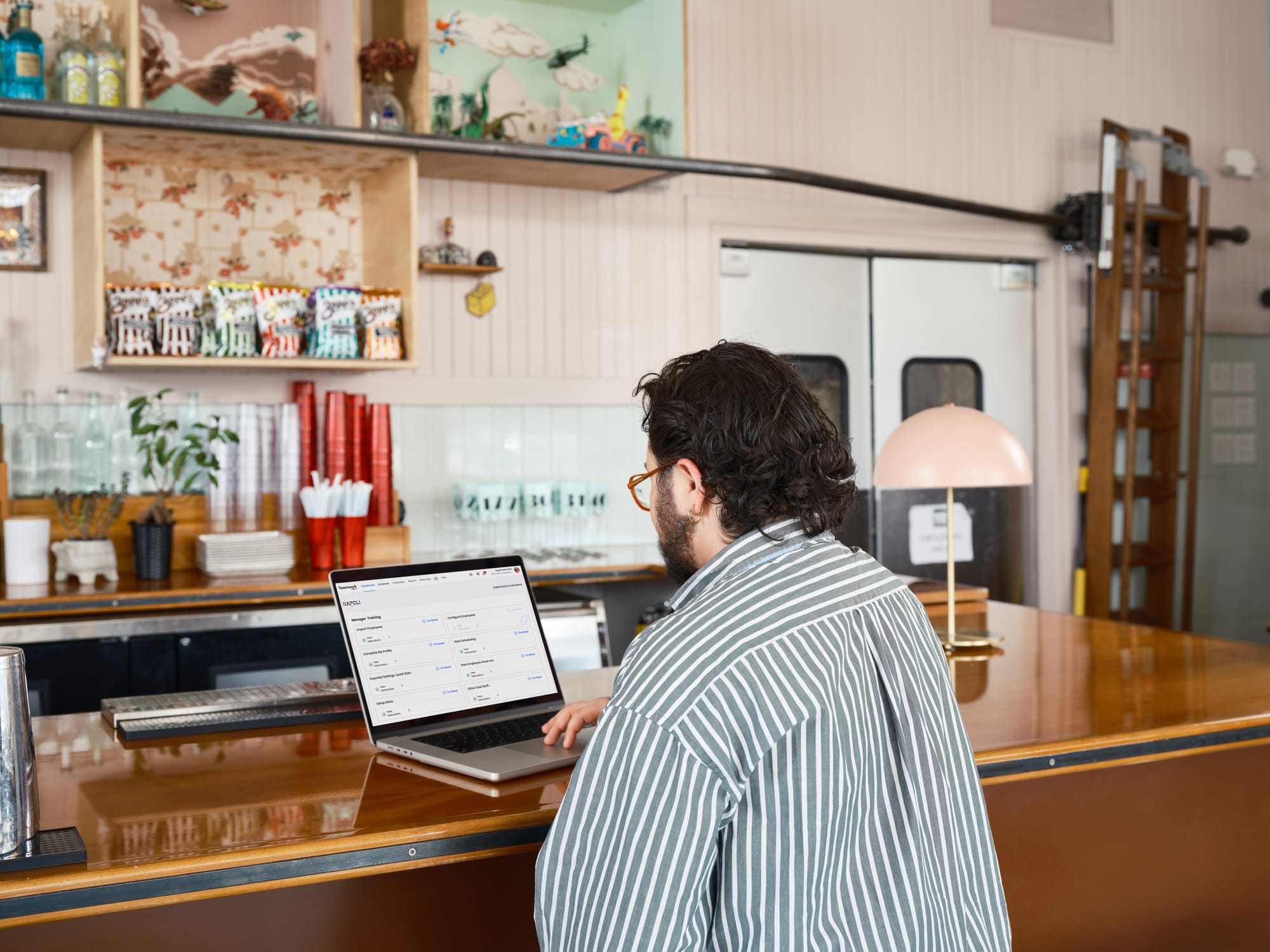 Restaurant manager using a laptop at a counter-service bar to review schedules and sales data while a bar staff member works in the background.