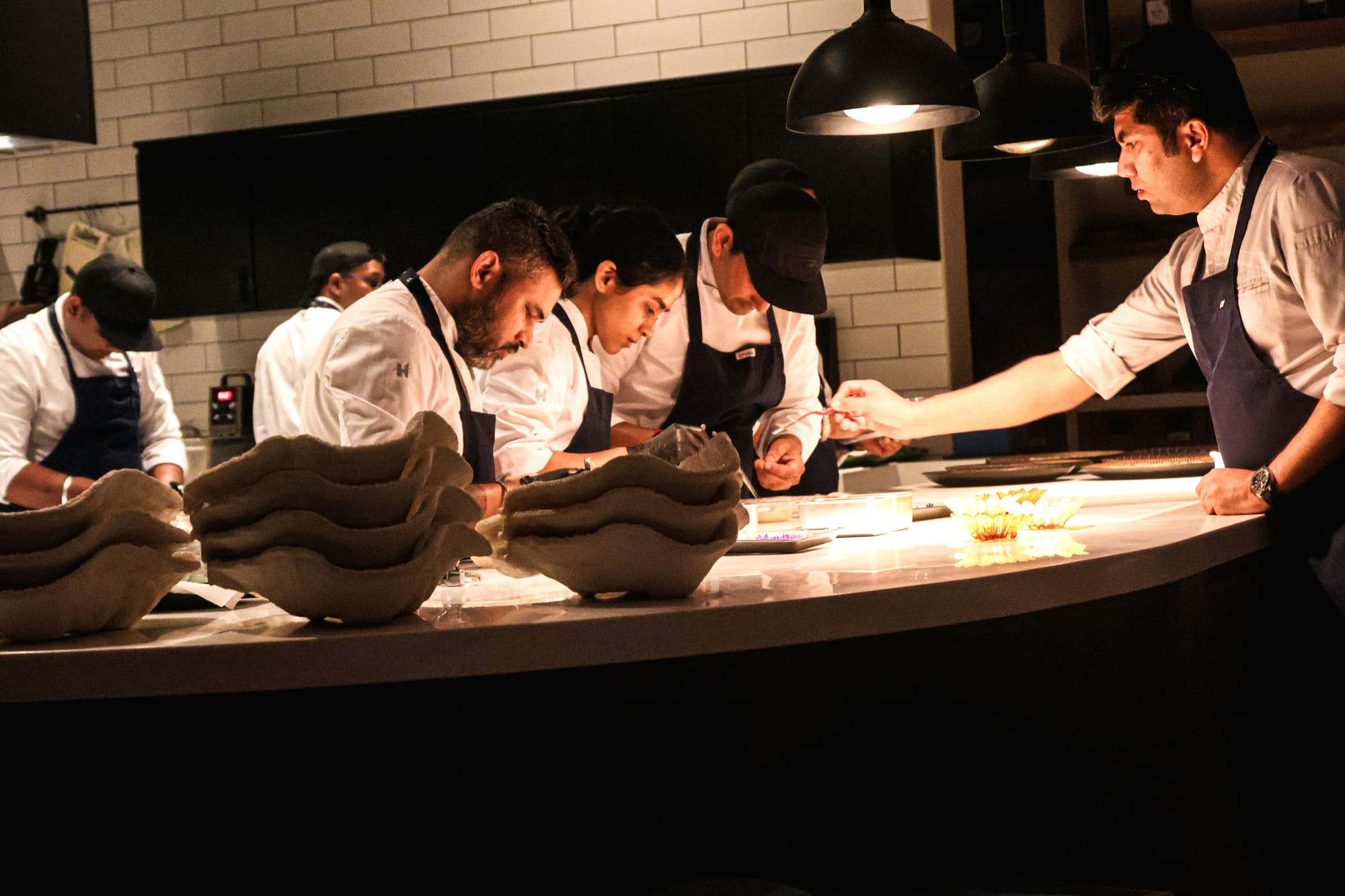 A row of chefs plates dishes together at a kitchen pass, with one team member handing off an ingredient to another.