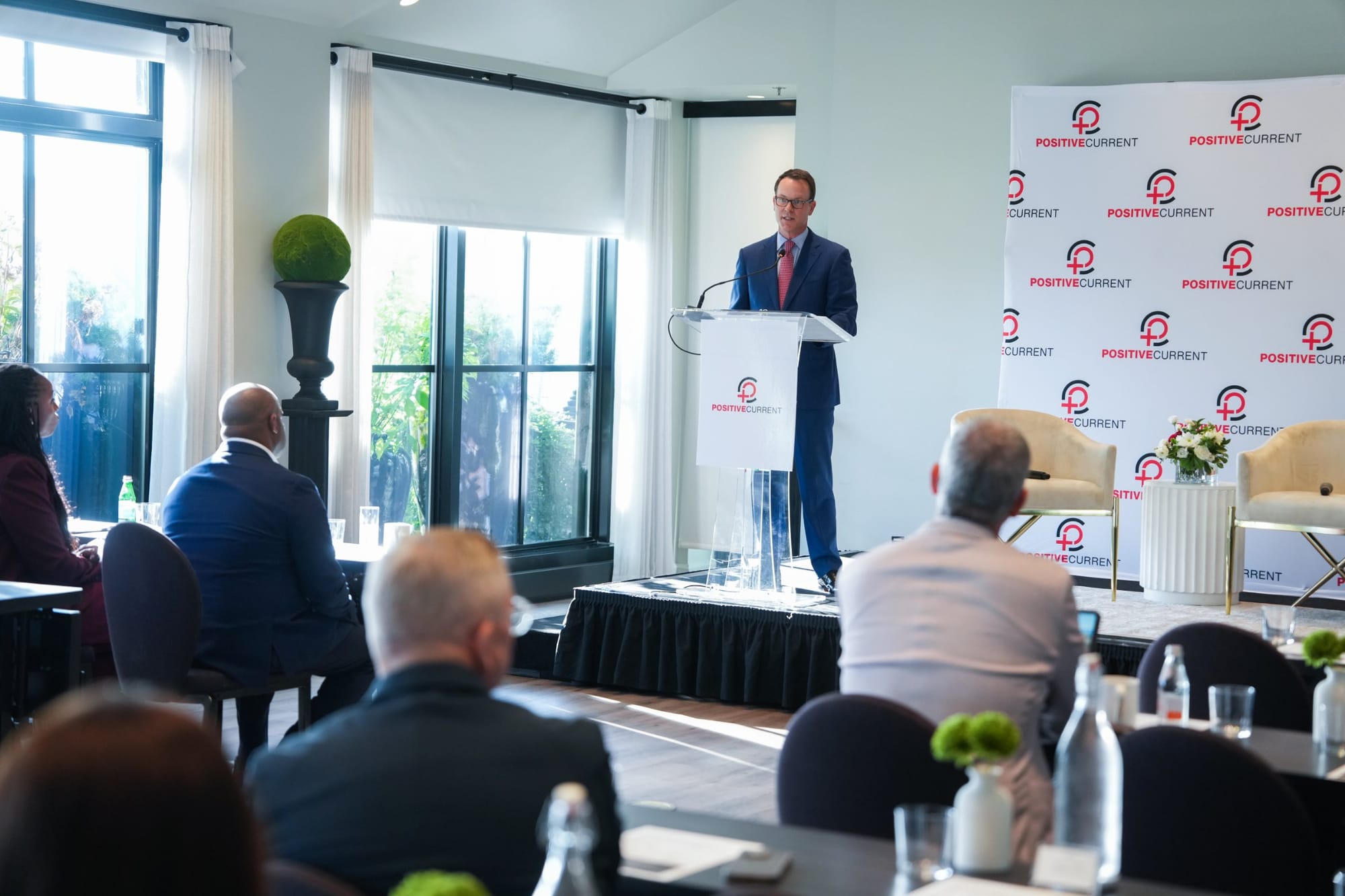 Amos Snead speaks at a podium in front of a Positive Current backdrop as guests listen from their tables