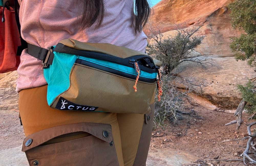a hiker wearing the 2.5L SwitchPack by Chicken Tramper Ultralight Gear demonstrates that a camera with a lens fits in the pack out on trail