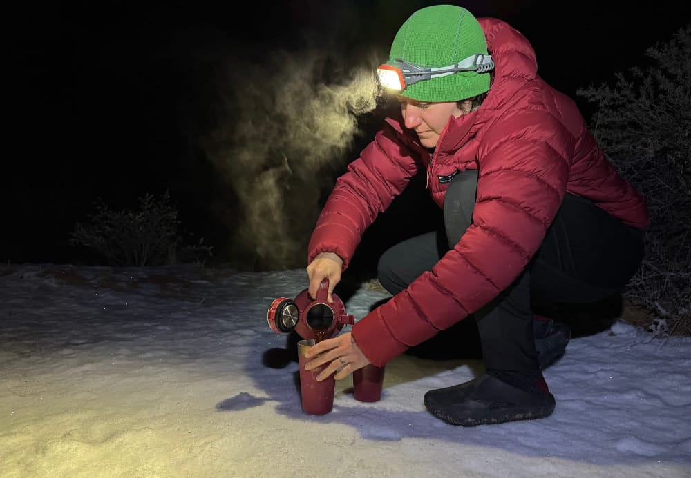 a camper pours hot coffee from the stanley easy-pour 640z growler in a snowy camp during winter with her headlamp on