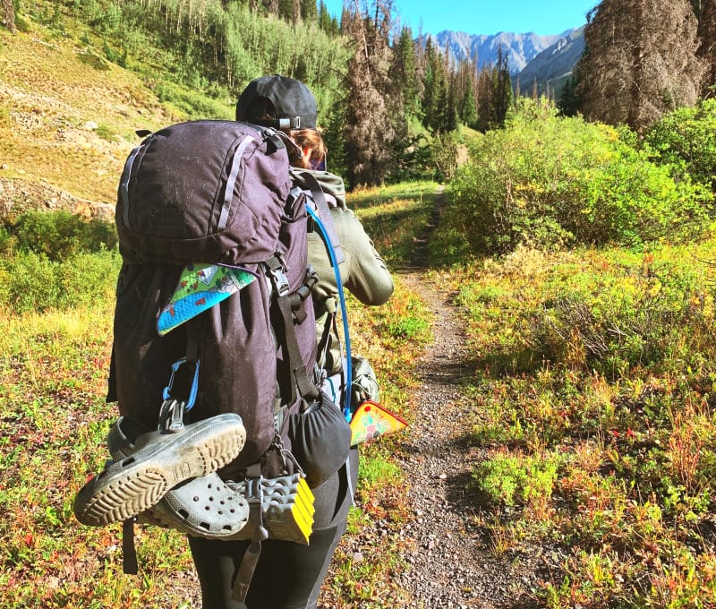 Backpacker hiking on a mountain trail with a Kula Cloth visibly clipped to their pack, emphasizing Leave No Trace outdoor hygiene.