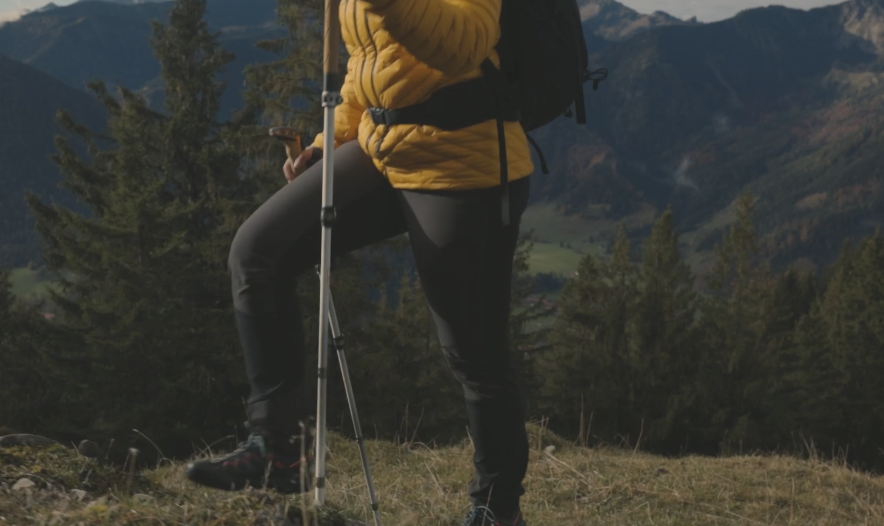  Woman hiker wearing a yellow jacket standing with LEKI Cressida FX Carbon AS trekking poles in a mountain landscape.