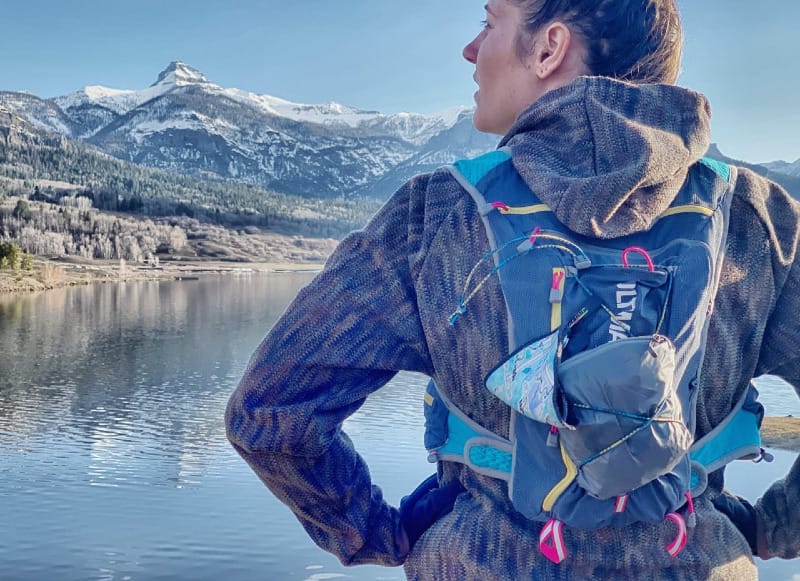  Female hiker standing near an alpine lake with snow-capped peaks in the background, wearing a running vest with a Kula Cloth attached.