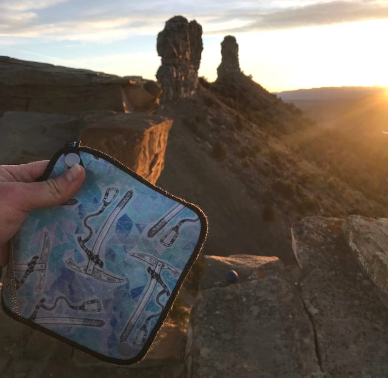 Hand holding a Kula Cloth with ice axe artwork during sunset on a rocky outcrop, with scenic desert spires in the background.