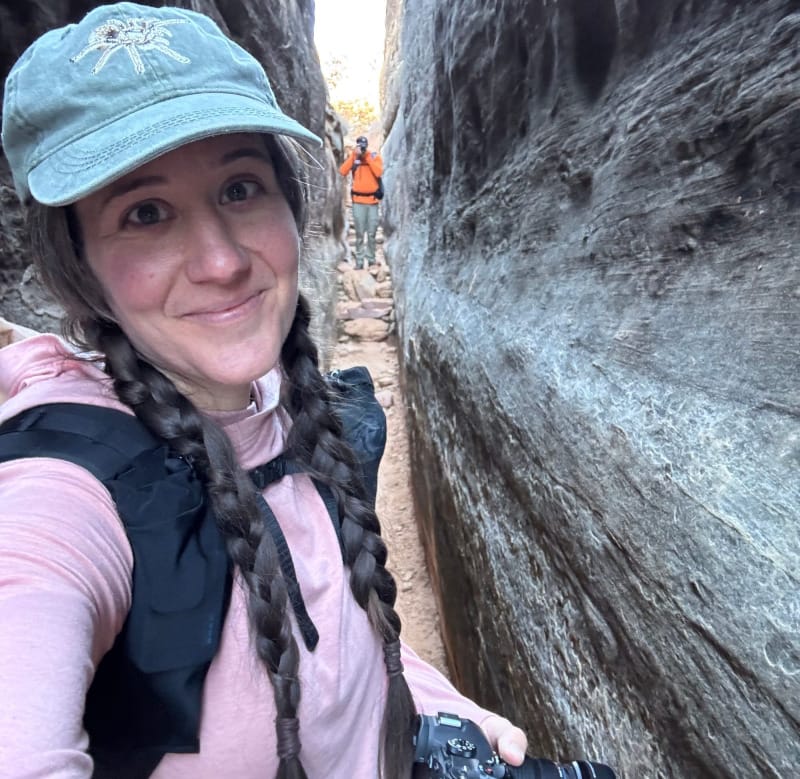 Hiker wearing a light pink sunshirt and braided pigtails, holding a camera and standing between narrow sandstone canyon walls.