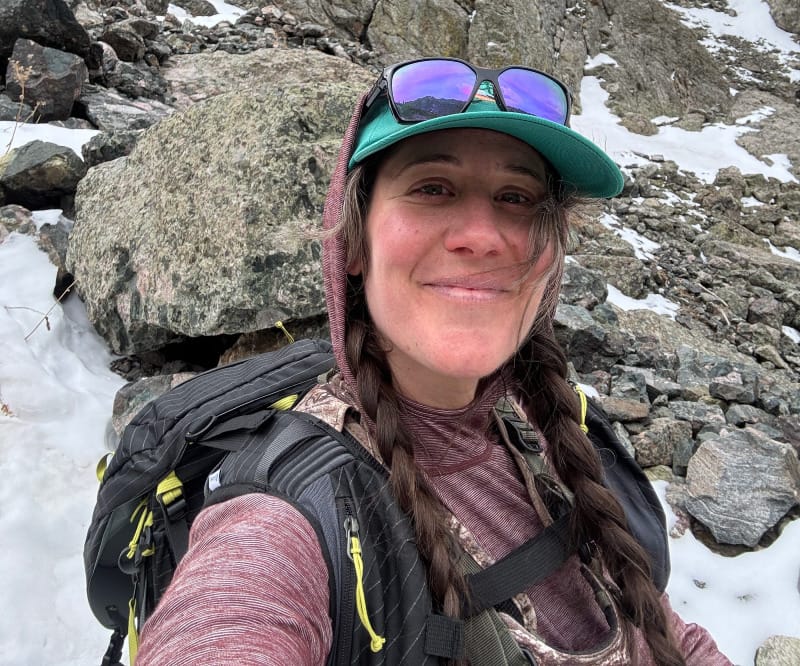 Backpacker smiling with the NEMO Persist 45L Backpack in steep, rocky alpine terrain with patches of snow.
