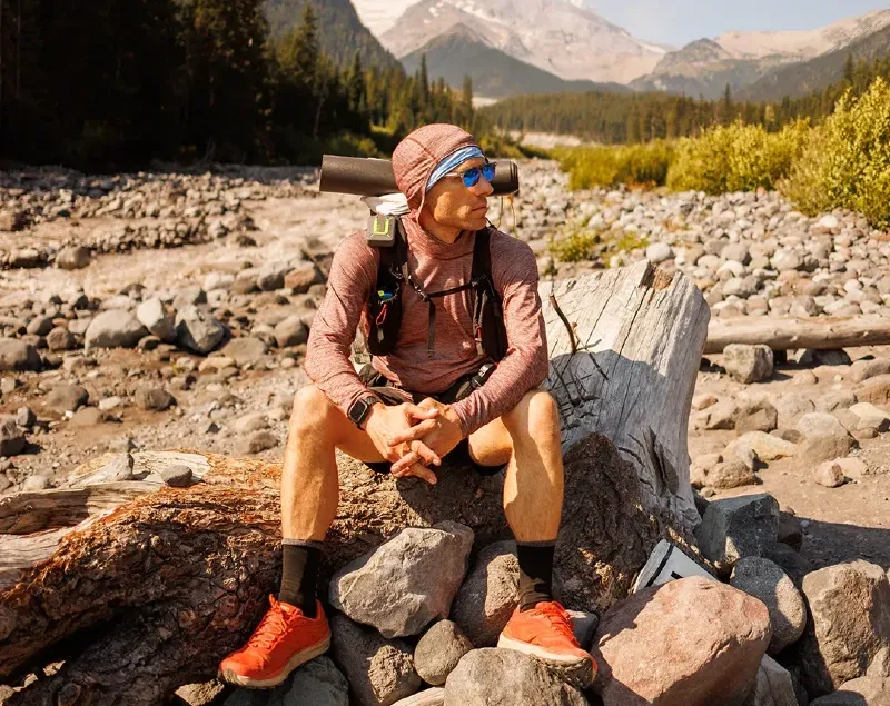 A hiker wearing the Tern Hoodie sits on a log with a mountainous landscape in the background.