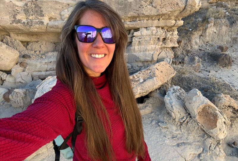 Woman smiling in a red fleece hiking shirt with a camera in a rocky desert landscape.