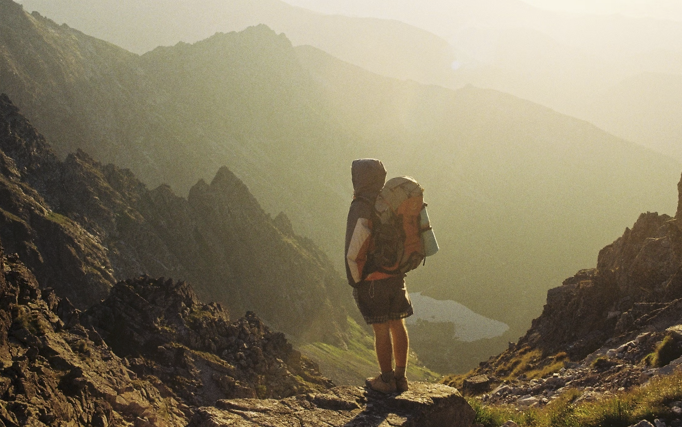 Backpacker wearing a hooded jacket and carrying a large pack stands on a rocky mountain ledge at sunrise, overlooking a deep alpine valley.