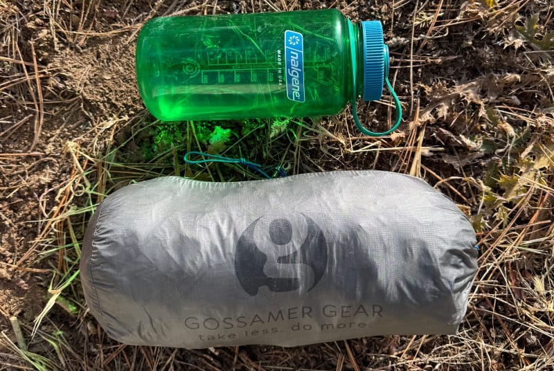 Gossamer Gear shelter packed next to a green Nalgene bottle on dry forest ground for size comparison.