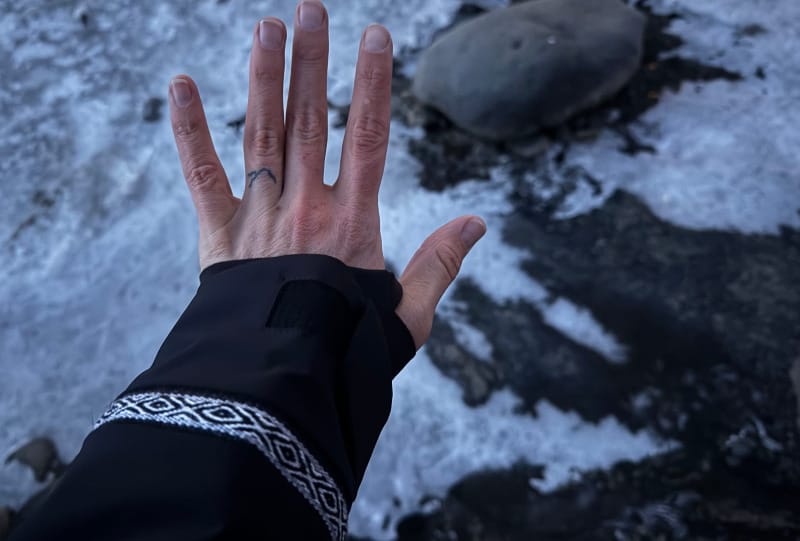 Hand showing signs of cold exposure over a frozen creek, wearing a black technical shell with snowflake embroidery.