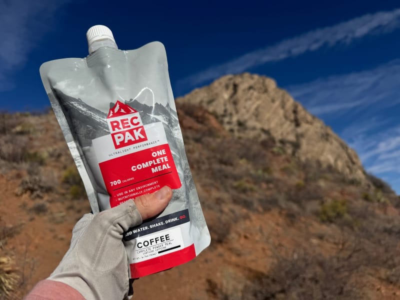  Gloved hand holding a RecPak Coffee meal pouch on a desert hiking trail with rocky terrain and a blue sky in the background.