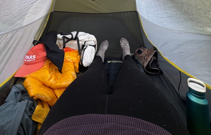 View from inside a tent with a person wearing Ibex leggings and surrounded by backpacking gear, including a puffy jacket, hat, and water bottle.