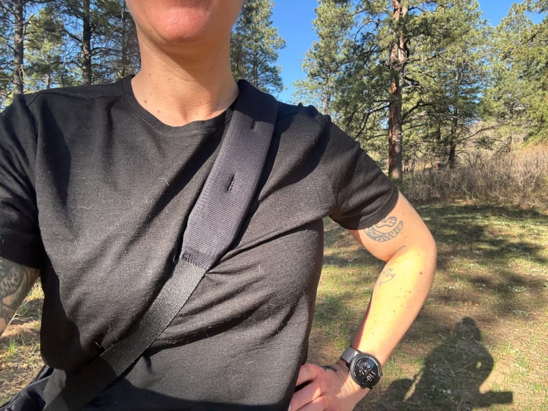 Person wearing the Ibex merino wool Go Far Tee in black, standing in a forested area with sunlight filtering through pine trees.