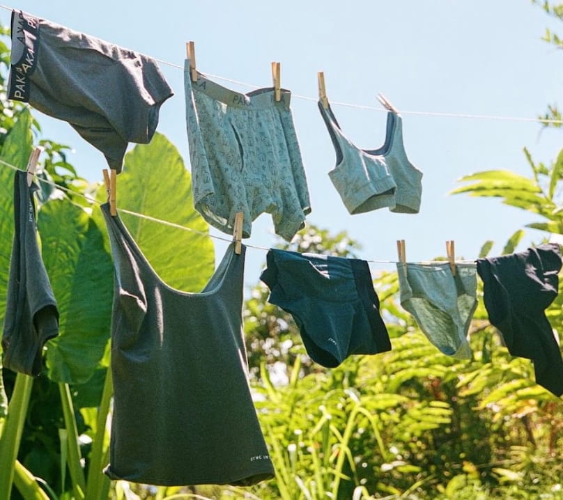  PAKA Essentials 2.0 underwear, bralettes, and tank tops hanging on a clothesline outdoors in a lush, tropical setting, with sunlight highlighting the soft fabric textures.