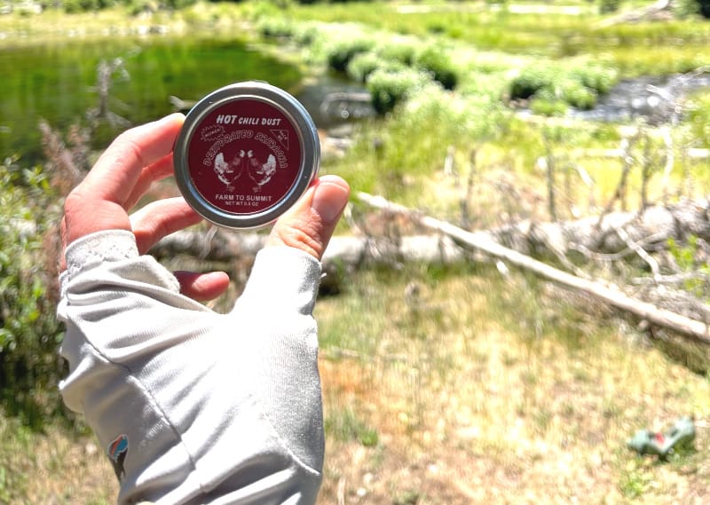  Hand holding a tin of Farm To Summit Hot Chili Dust with a dramatic mountain range and alpine meadow in the background.