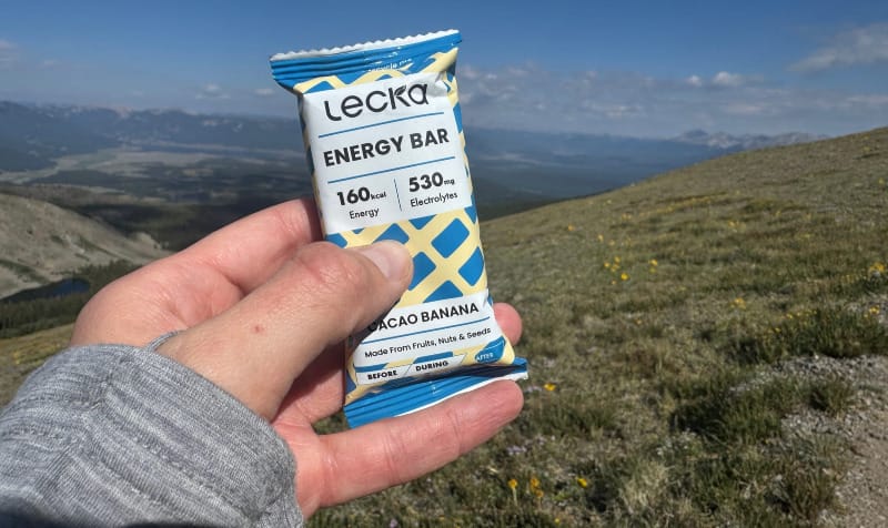 A hand holds a Lecka Cacao Banana Energy Bar on a high mountain ridge with scenic alpine views in the background.