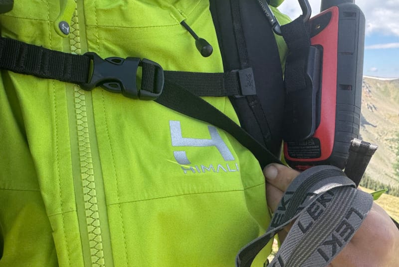  Close-up of HIMALI Monsoon Jacket chest area showing logo, waterproof zipper, and backpack chest strap with GPS device attached.