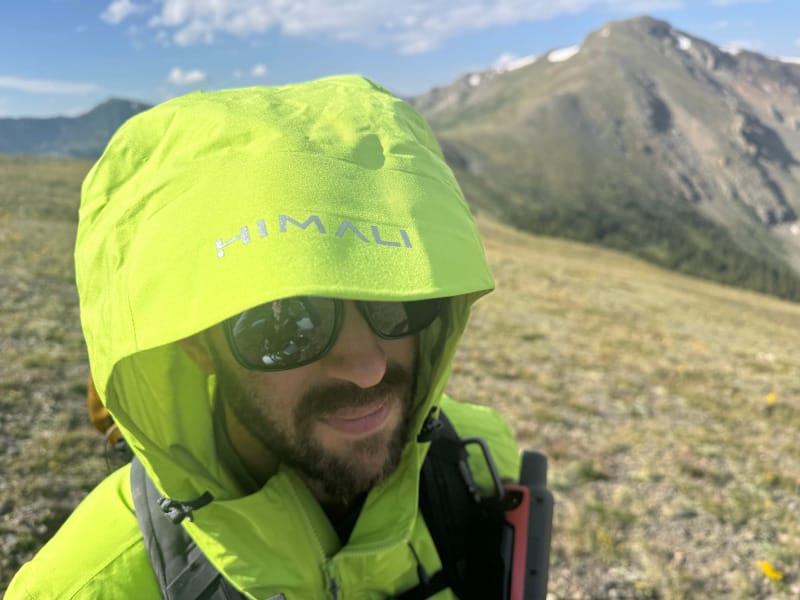  Zoomed-in view of a hiker wearing the HIMALI Monsoon Jacket with hood up, sunglasses on, and alpine peaks in the background.
