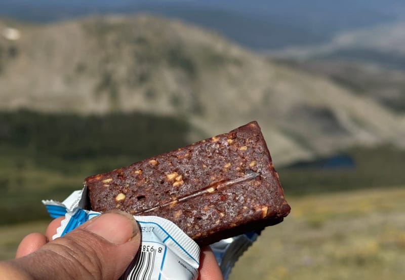 A close-up of a bitten Lecka Cacao Banana Energy Bar held up with a mountain backdrop in the distance.