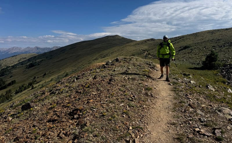  Backpacker wearing a bright green HIMALI Monsoon Jacket hiking along an exposed ridgeline under a dramatic sky in the mountains.