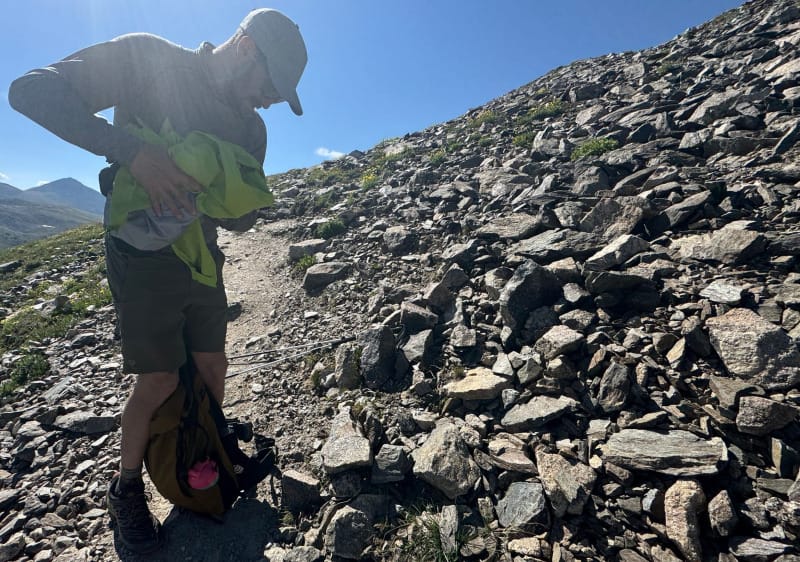  Hiker on a rocky alpine trail stuffing a neon green HIMALI Monsoon Hardshell Jacket into their backpack under clear blue skies.