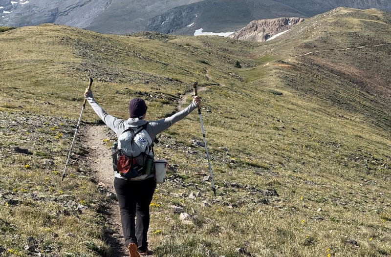 Hiker raising trekking poles while wearing the new Windrider 40 pack, hiking through alpine meadows.