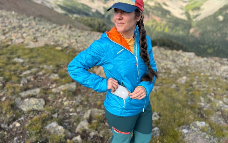 Woman hiking in the Mammut Eigerjoch Light IN Hooded Jacket, reaching into the zippered pocket with rugged alpine terrain in the background.
