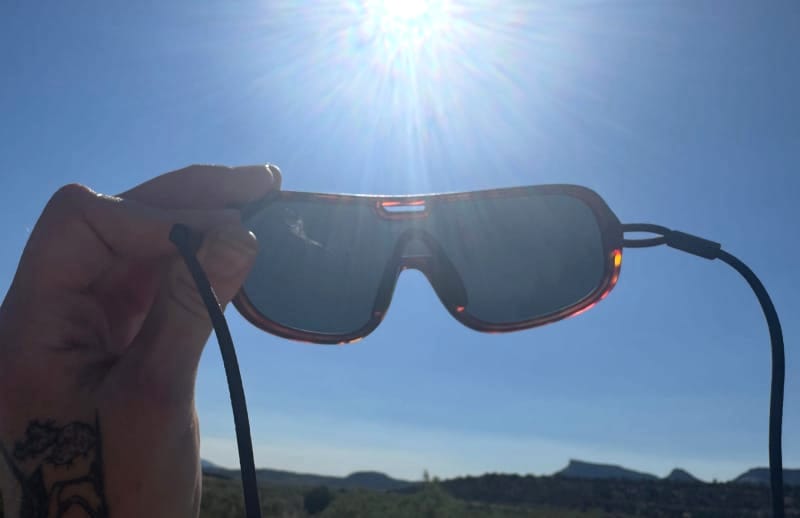  Hand holding up Ombraz Refugio sunglasses against the bright sun and blue sky.