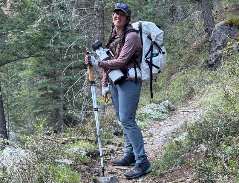 Backpacker standing on a rocky trail in the forest with trekking poles and a loaded hiking pack.
