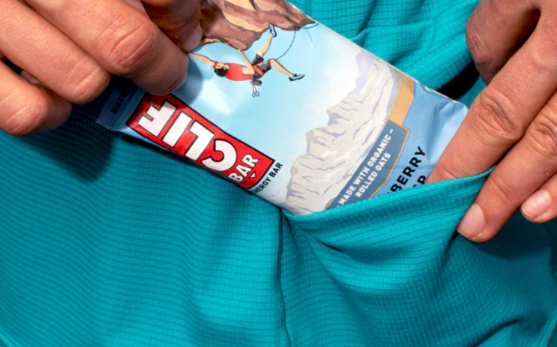 HIMALI Eclipse Sun Hoodie kangaroo pocket holding a Clif Bar, highlighting storage convenience for hikers.