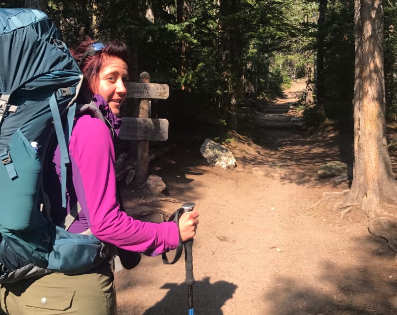 Backpacker standing at a wooden trail sign with a large hiking backpack and trekking pole.