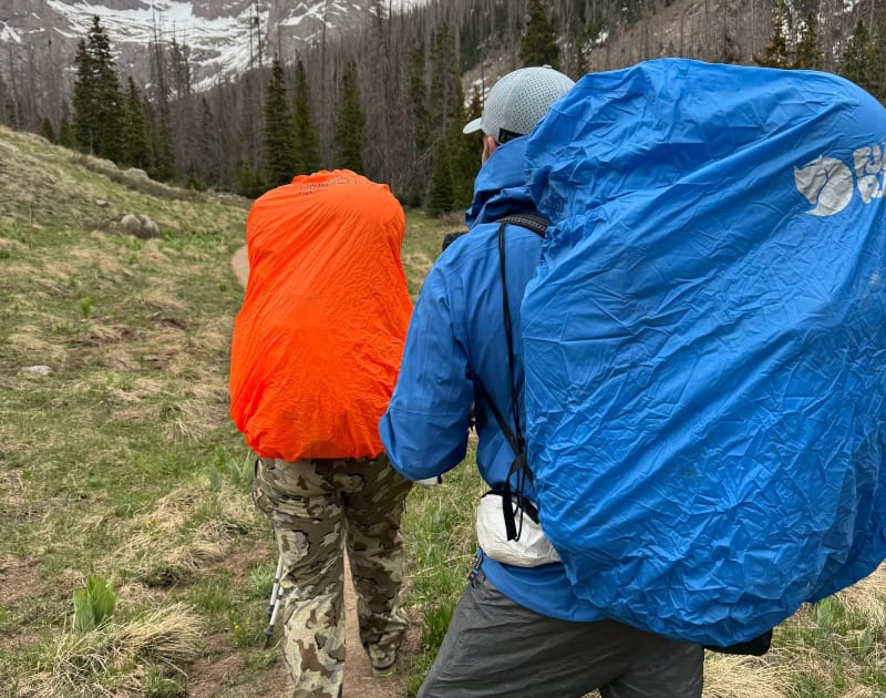 two backpackers hiking in the mountains with rain covers on their backpacks.