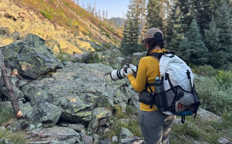Hiker in KUIU Tiburon Pants carrying a camera, climbing rocky terrain surrounded by pine trees.